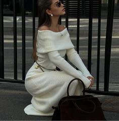 Woman in a white off-shoulder sweater and skirt sitting on a street corner.