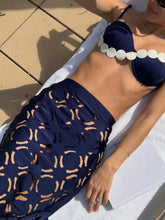 Close-up of a person wearing a blue bikini top with white scalloped trim and a matching blue maxi skirt with intricate geometric cut-outs, lying on a white towel in the sun.