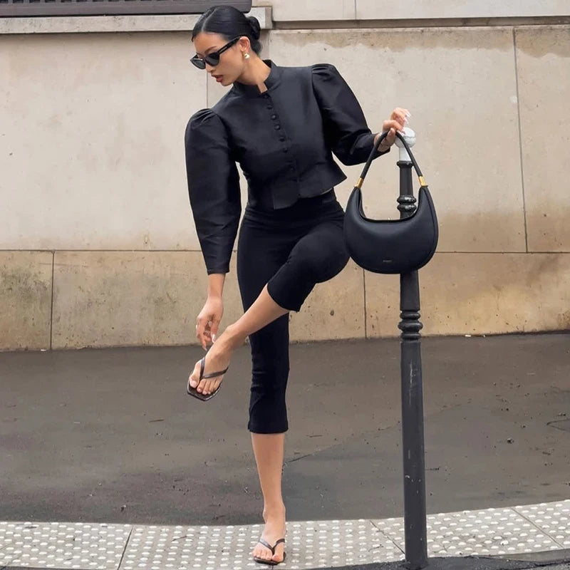 Woman in a black outfit posing with a black handbag against a beige wall.