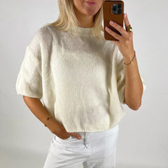 Person wearing a cream sweater and white pants, taking a mirror selfie.
