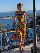 Woman in a colorful floral dress standing on a balcony with a scenic view of water and mountains.