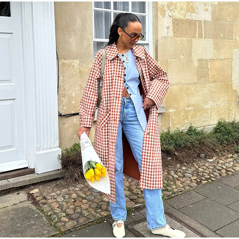 Woman in a checkered coat holding yellow flowers, standing on a sidewalk.