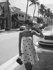 Woman in a polka dot dress taking a selfie on a street with palm trees and cars in the background.