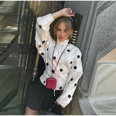 Woman wearing a white jacket with black polka dots, standing in front of a building entrance.