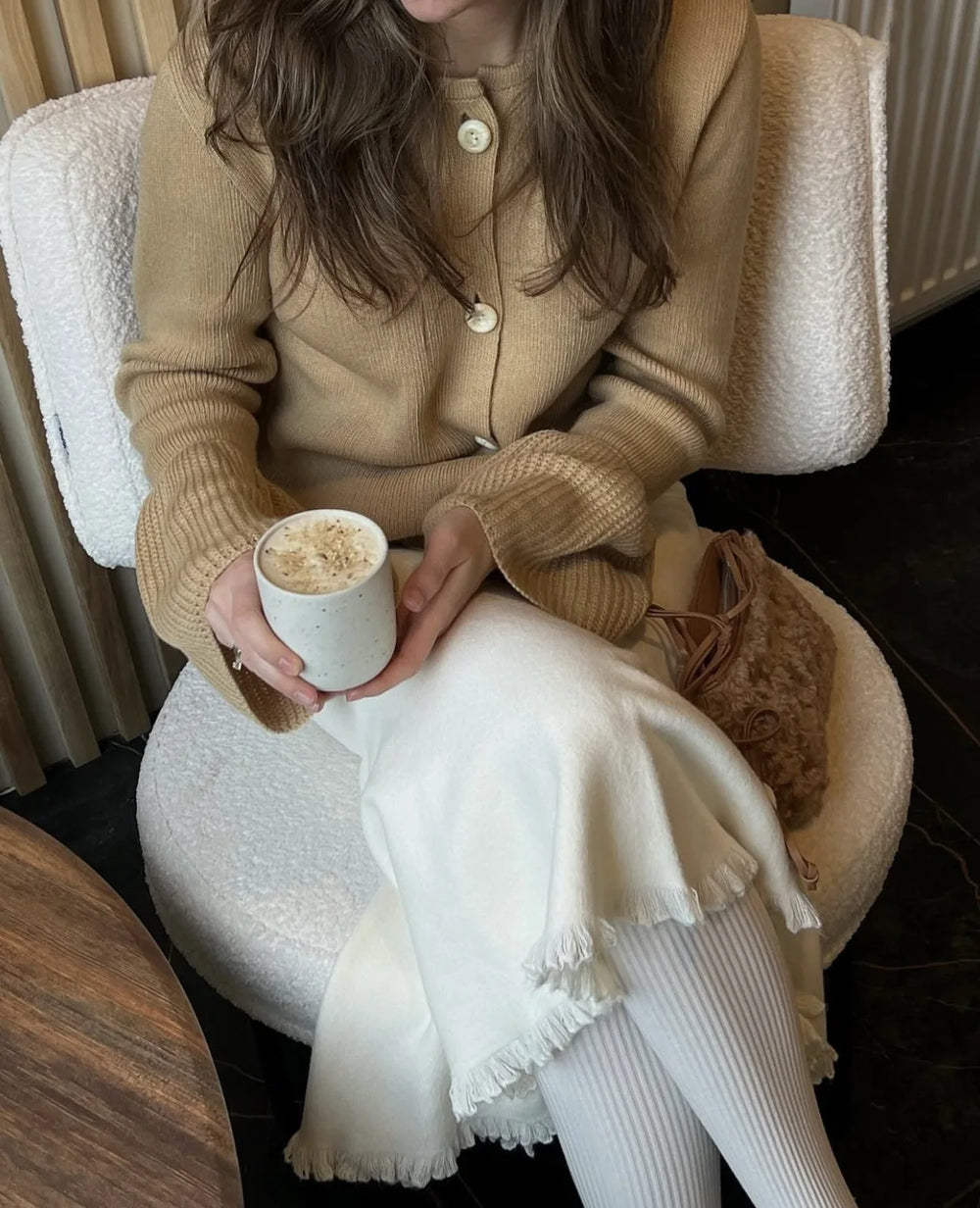 Person sitting in a chair holding a cup of coffee, wearing a beige coat and white pants.