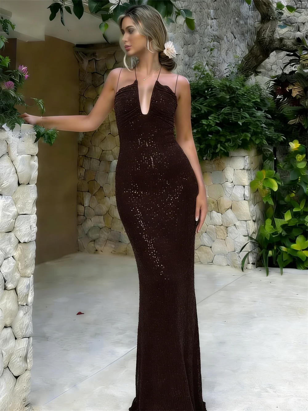 Woman in a dark brown evening gown standing outdoors with stone wall and greenery.