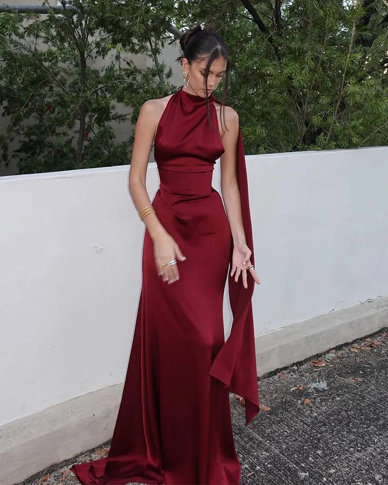 Woman wearing a burgundy evening gown with a halter neck and cape against a white wall and greenery background.