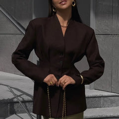 Woman wearing a dark brown blazer with a gold chain belt against a gray wall.