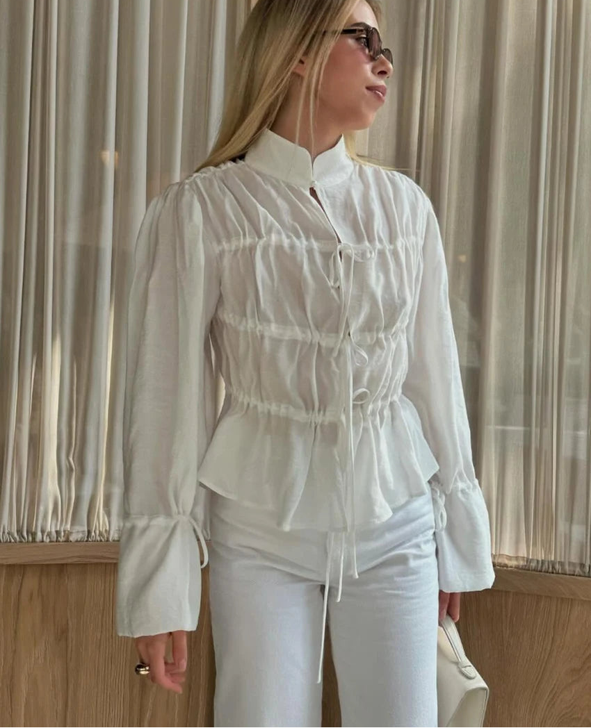 Woman wearing a white blouse with ruffled details and white pants, standing indoors.