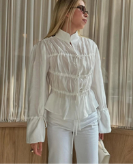 Woman wearing a white blouse with ruffled details and white pants, standing indoors.