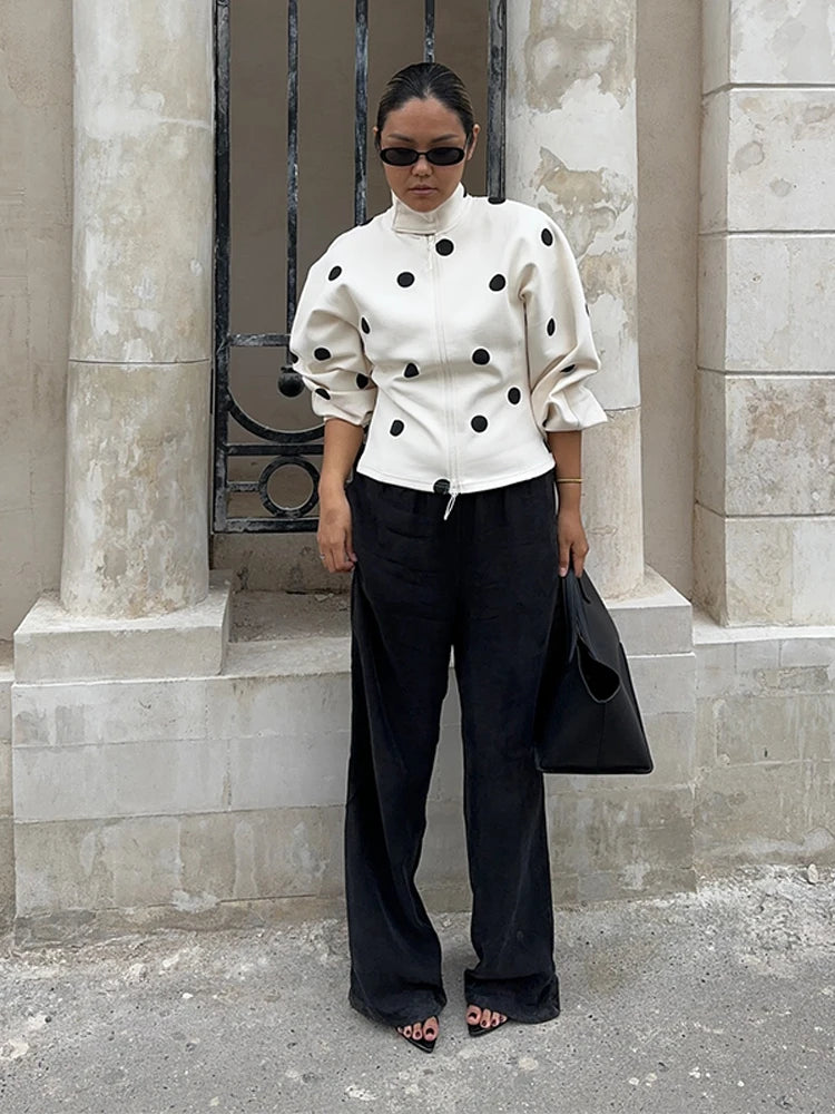 Person wearing a white polka dot blouse and black pants standing against a stone wall.