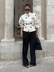 Person wearing a white polka dot blouse and black pants standing against a stone wall.