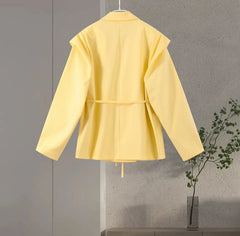 Yellow coat on a hanger against a gray wall with a plant in the corner.