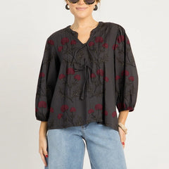Woman wearing a black blouse with red floral embroidery and blue jeans on a light gray background