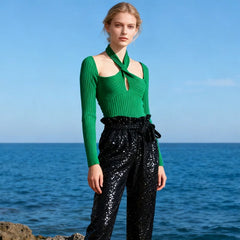 Woman wearing a green top and black sequin pants standing by the ocean.