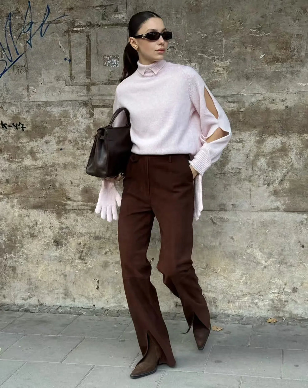 Woman in a light pink blouse and brown pants standing against a textured wall.