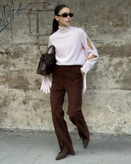 Woman in a light pink blouse and brown pants standing against a textured wall.
