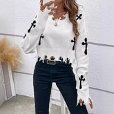 A white sweater with black cross patterns worn by a person against a white wall.