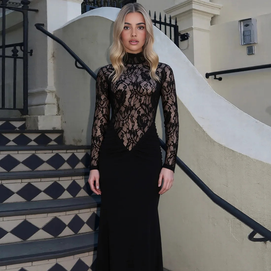 Woman in a black lace dress standing on a staircase with a white wall and black railing in the background.