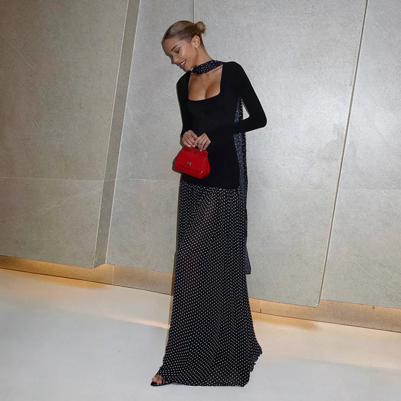 Woman in a black long-sleeve top and polka dot skirt holding a red handbag against a neutral wall.