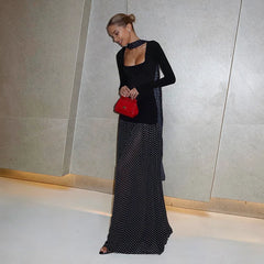 Woman in a black long-sleeve top and polka dot skirt holding a red handbag against a neutral wall.