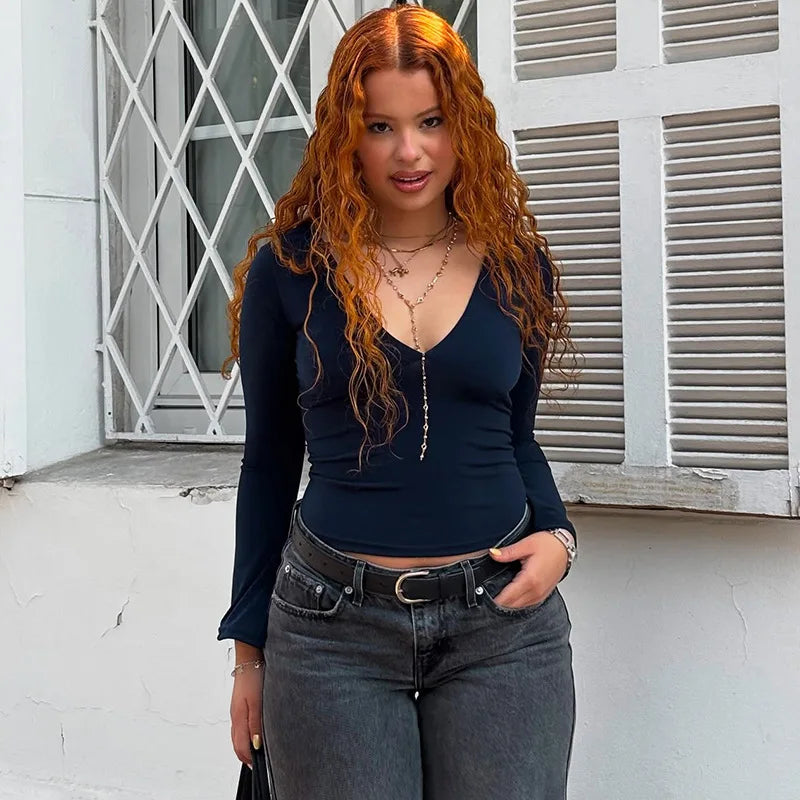 Woman with long, wavy hair wearing a dark blue top and gray jeans standing in front of a white wall.