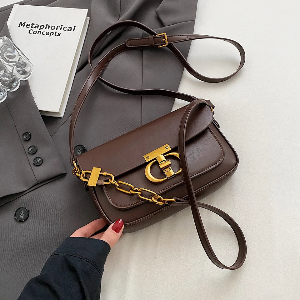 Brown handbag with gold chain detail held by a hand on a light surface with a gray coat and book in the background.