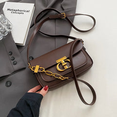 Brown handbag with gold chain detail held by a hand on a light surface with a gray coat and book in the background.