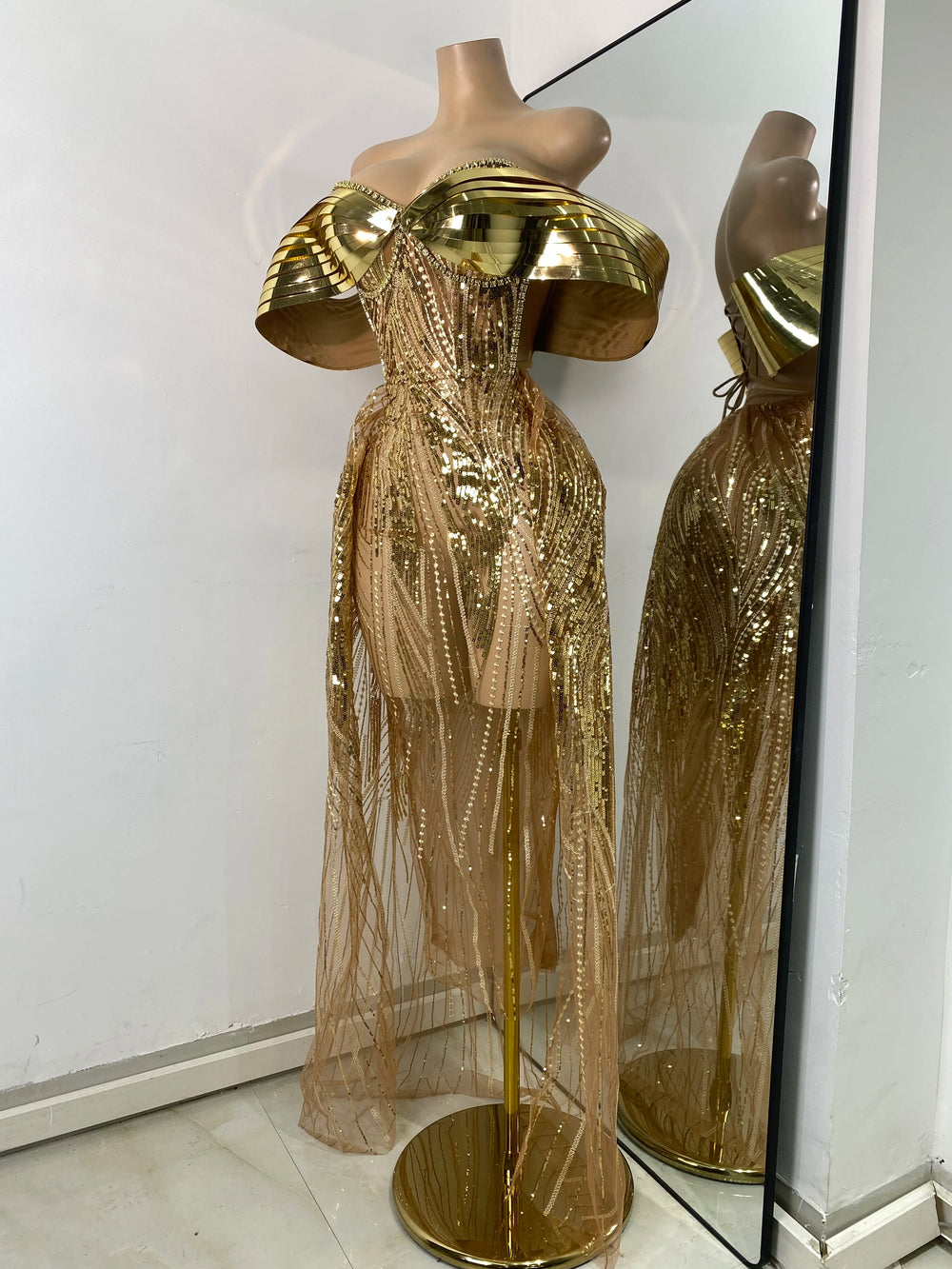 Gold sequin dress on a mannequin with a mirror reflection against a white wall.