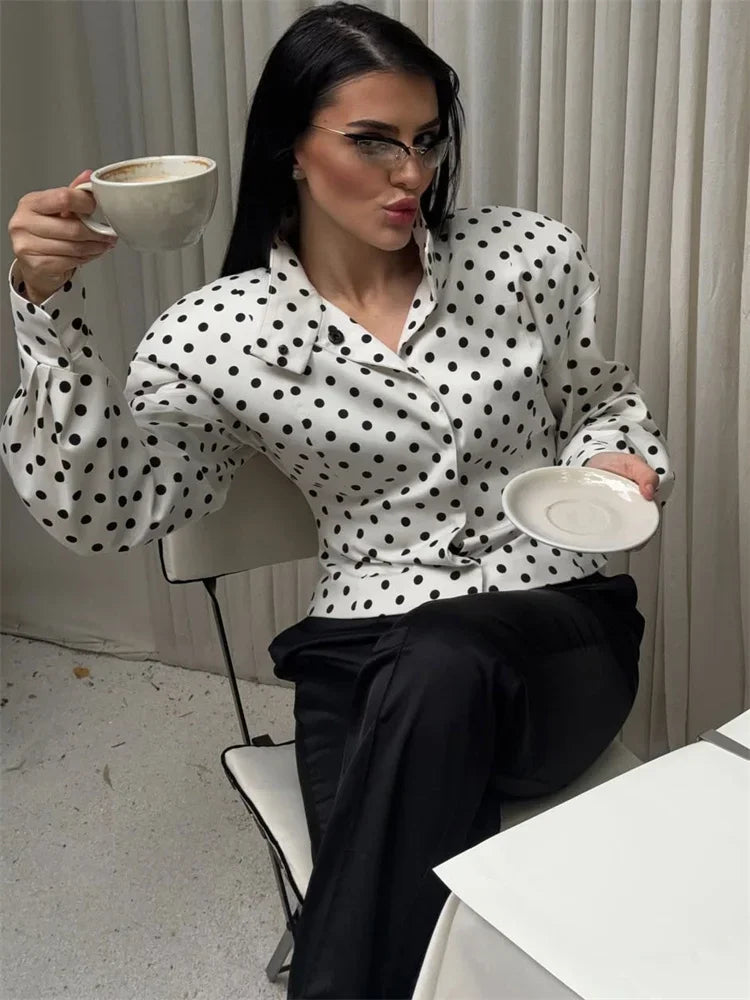 Woman in a polka dot blouse holding a cup and saucer in a minimalistic room.