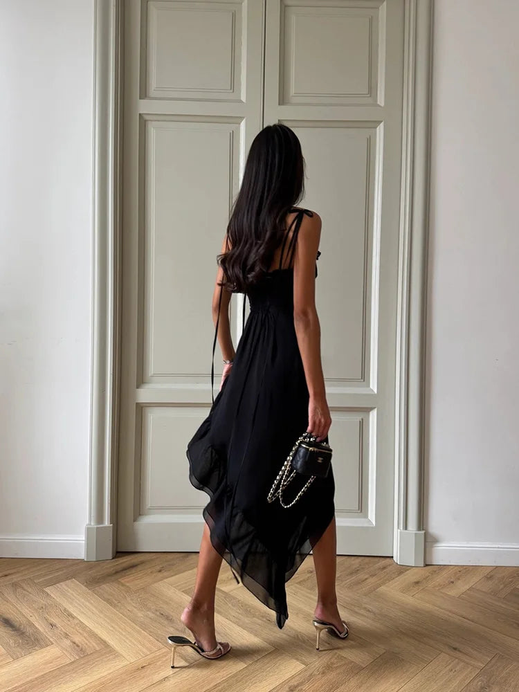 Woman in a black dress standing in a room with wooden flooring and white walls.