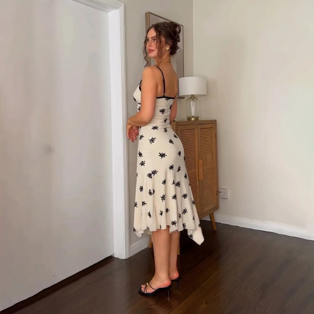 Woman in a white dress with black patterns standing in a room with a lamp and wooden cabinet.