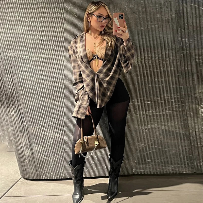 Woman taking a mirror selfie wearing a plaid shirt, black leggings, and high boots against a textured wall.