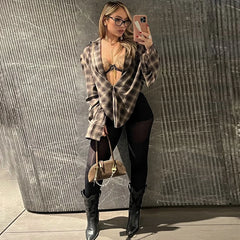 Woman taking a mirror selfie wearing a plaid shirt, black leggings, and high boots against a textured wall.