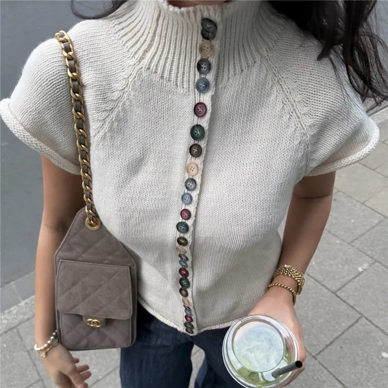 Person wearing a light gray sweater with colorful buttons, holding a brown handbag and a drink.