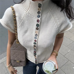 Person wearing a light gray sweater with colorful buttons, holding a brown handbag and a drink.