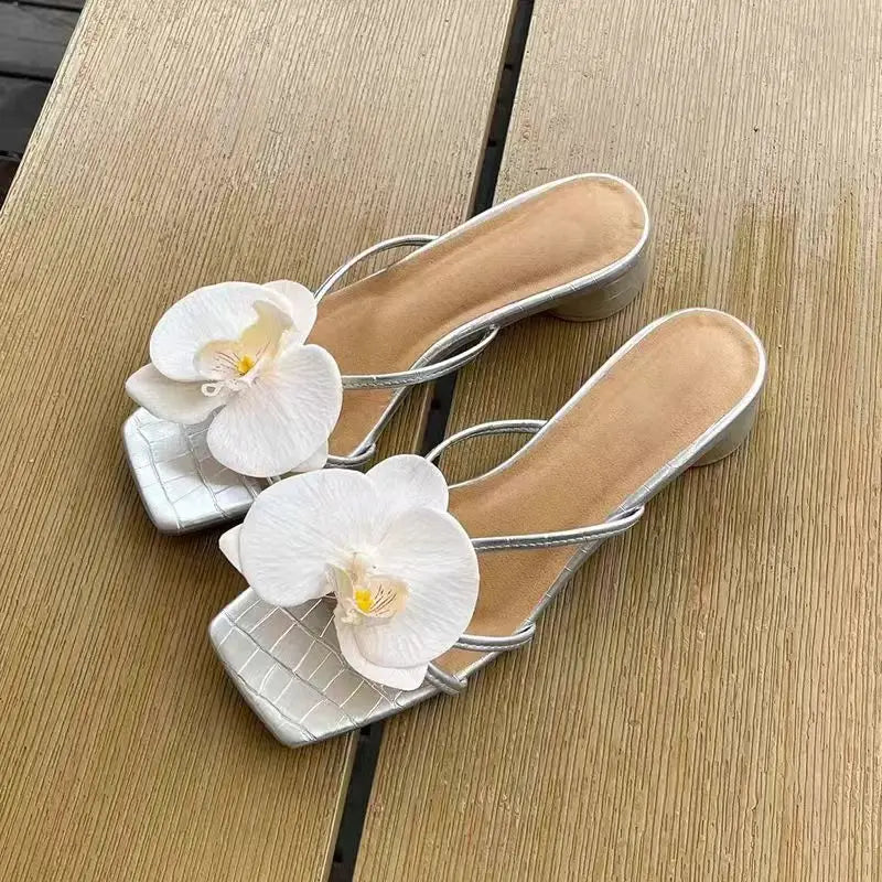 Silver high-heeled sandals with white floral decorations on a wooden surface