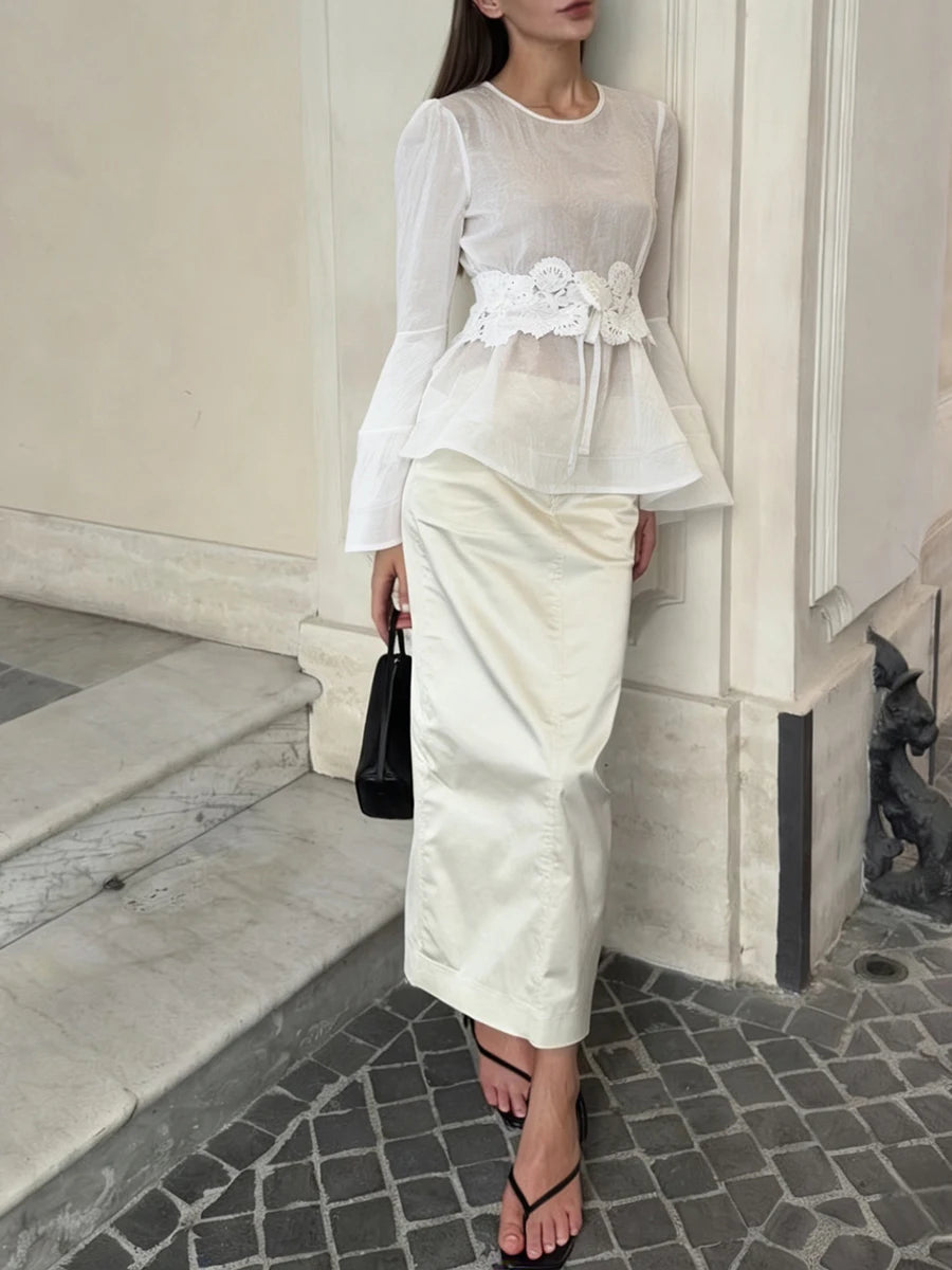 Woman in a white outfit with floral details standing against a neutral wall.
