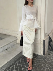 Woman in a white outfit with floral details standing against a neutral wall.