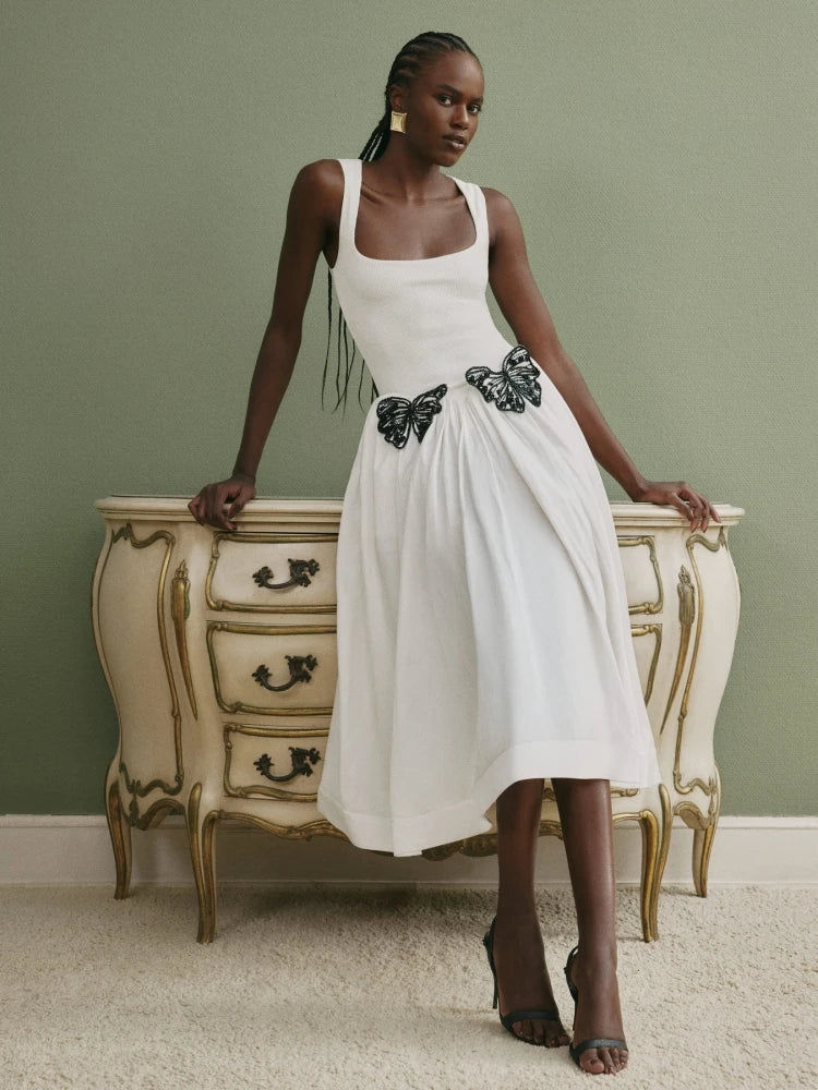 Woman in a white dress with butterfly embellishments standing next to an ornate dresser against a green wall.