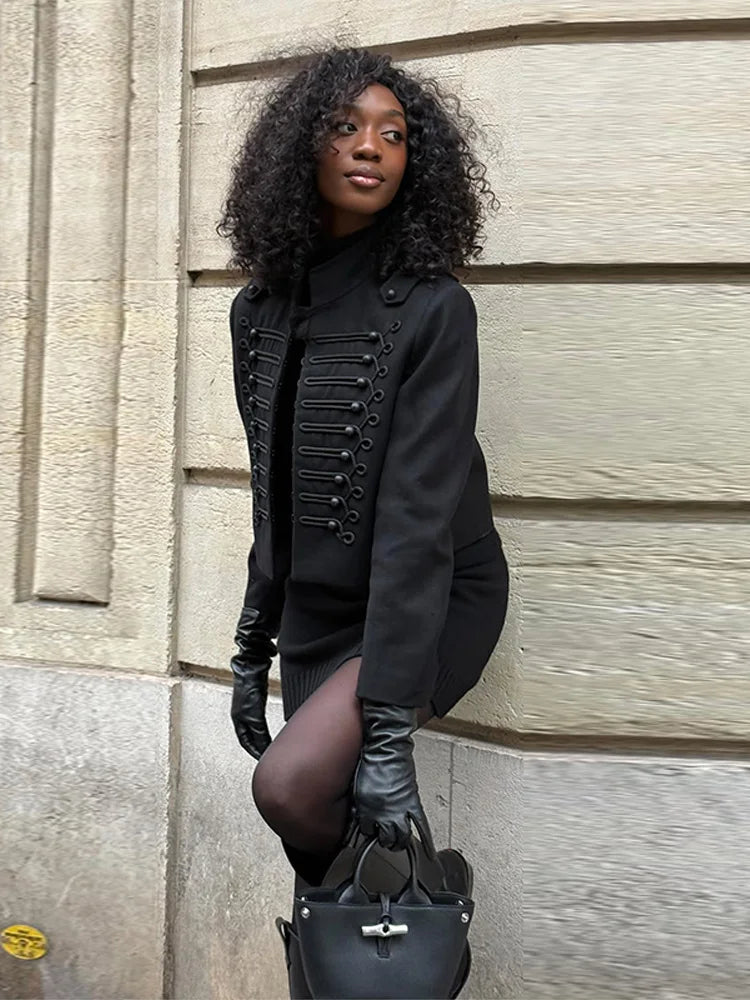 Woman in black coat and gloves standing against a textured wall.