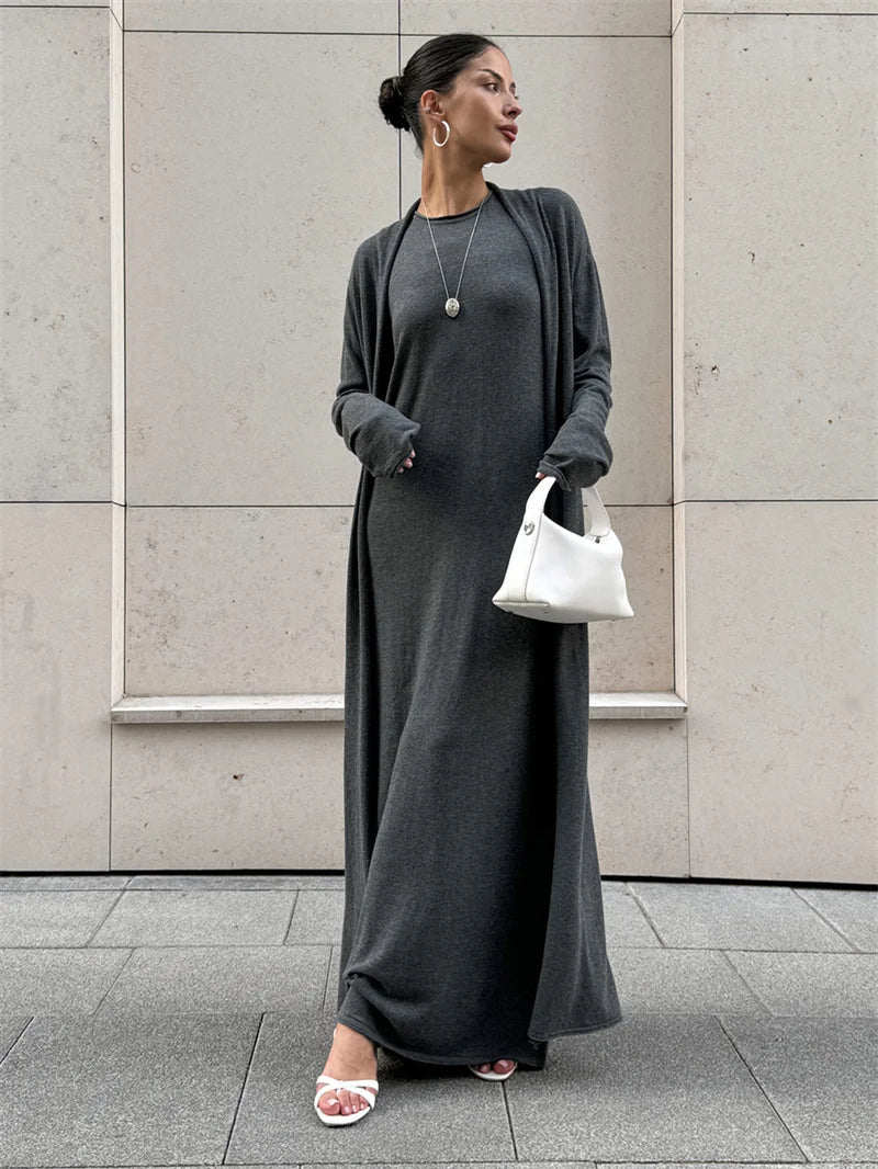 Woman wearing a long gray dress with a white handbag against a light-colored wall.