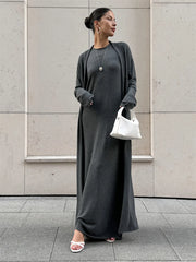Woman wearing a long gray dress with a white handbag against a light-colored wall.