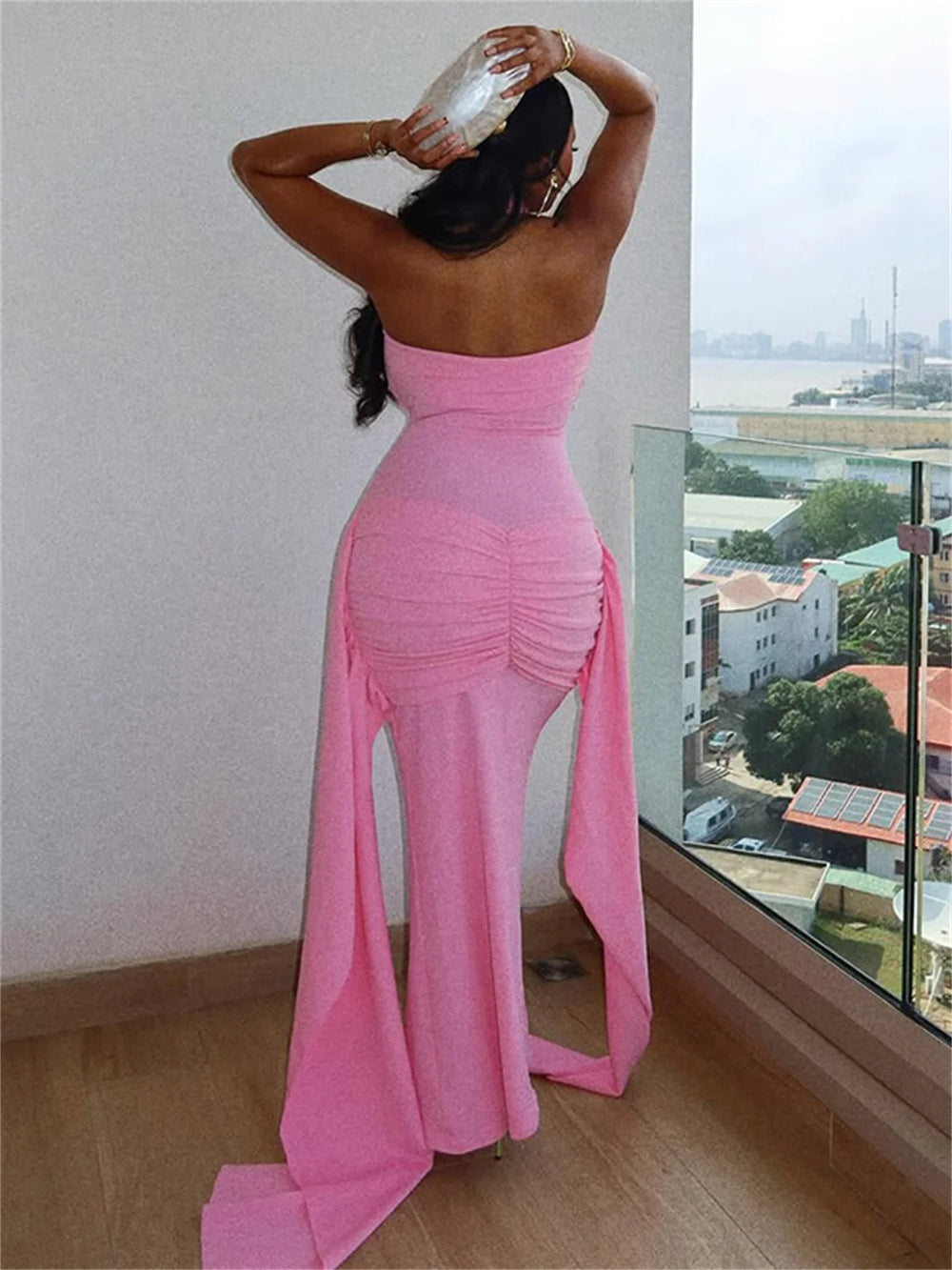 Woman in a pink strapless dress standing by a window with a cityscape view.