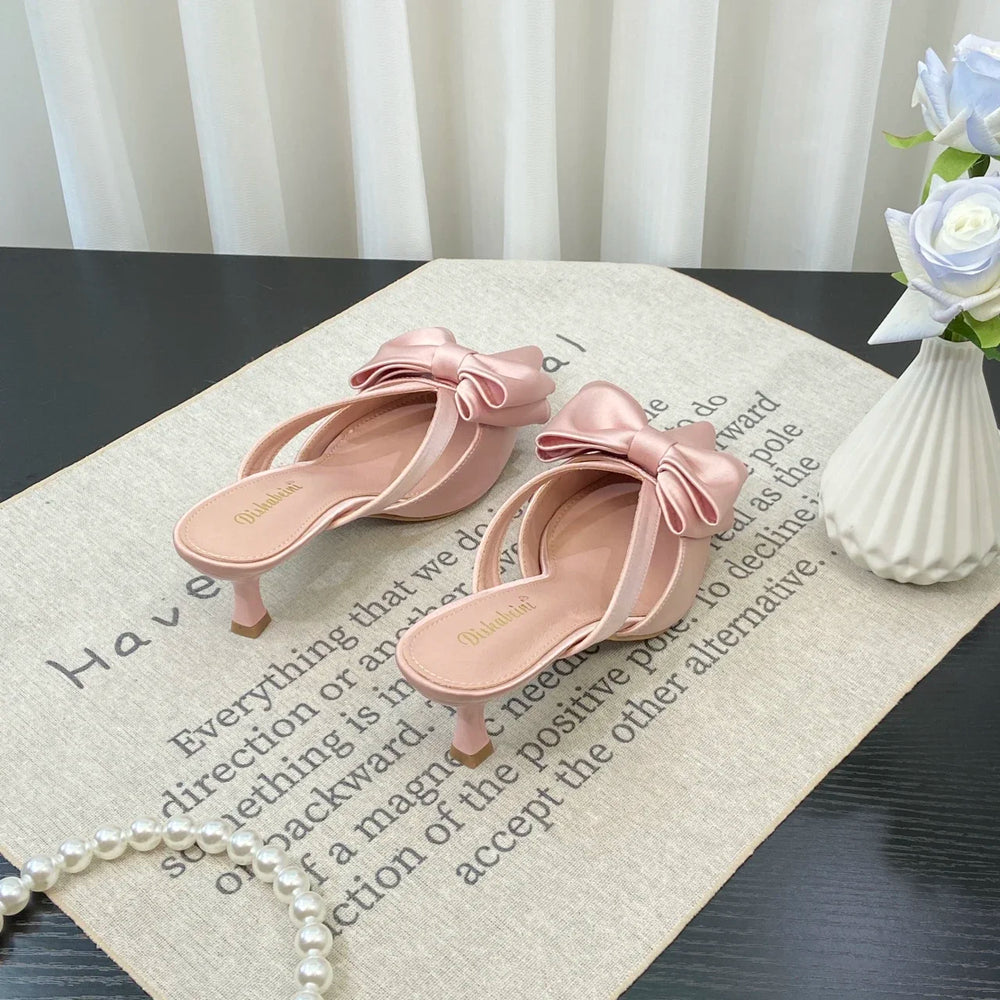 Pink high-heeled sandals with bow details on a textured surface with a vase of flowers in the background.