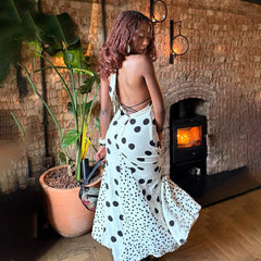 Woman in a polka dot dress standing in front of a lit fireplace with plants around.
