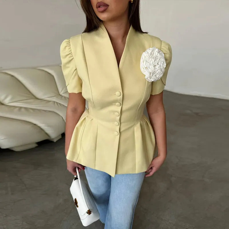 Woman wearing a light yellow blazer with a floral detail, holding a white handbag indoors.