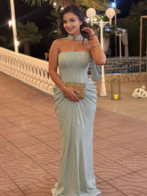 Woman in a light blue strapless dress holding a gold clutch on a paved walkway at night.