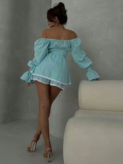 Woman wearing a light blue off-shoulder dress with puffed sleeves against a gray wall.