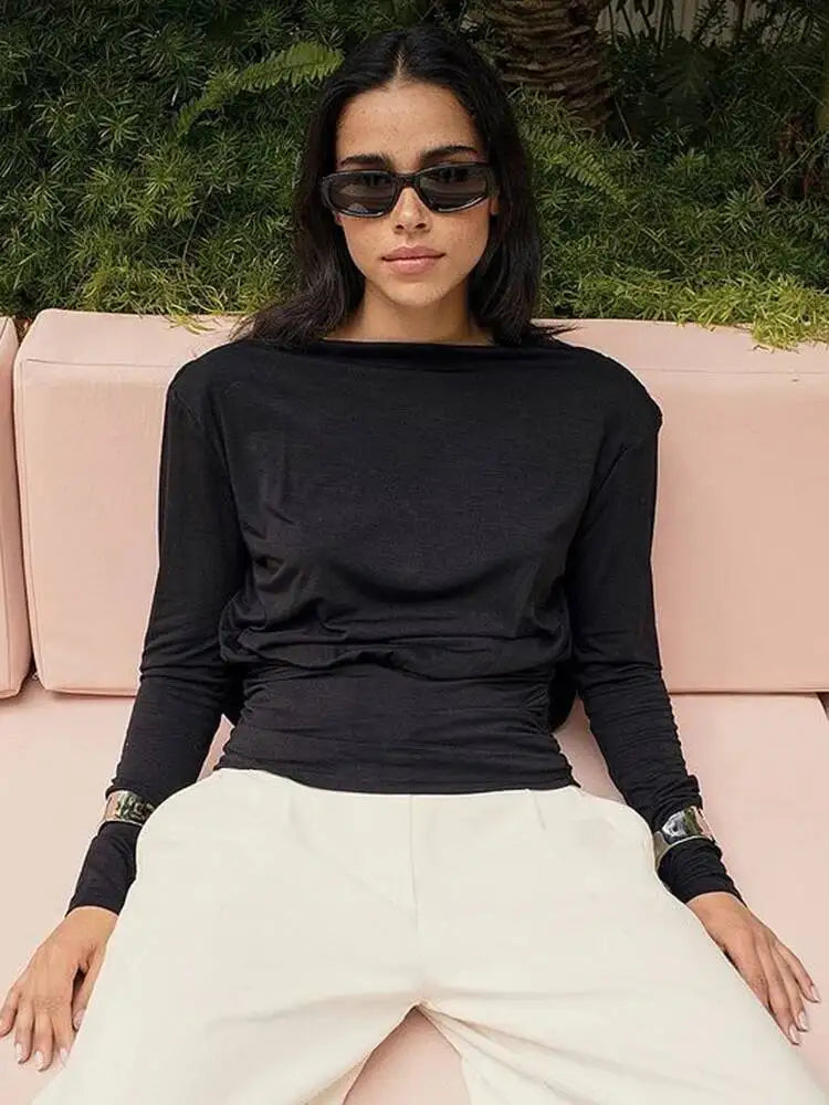 Woman wearing a black long-sleeve top and white pants sitting on a pink couch with greenery in the background.